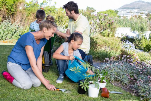 A family engaged in gardening activities in a well-maintained backyard garden, featuring a lush green lawn with evenly cut grass, bordered by flower beds filled with blooming plants and shrubs. The foreground shows a woman kneeling on the grass, planting or tending to a small shrub while holding a gardening fork, with gardening tools and potted plants nearby. Two children are assisting, one holding a watering can, and another sitting on the grass, actively participating. Behind them, a man and woman stand together, smiling and enjoying the outdoor space. The garden has a mix of cultivated flower beds and natural foliage, with a paved patio area visible in the background. The scene is bathed in bright sunlight under a clear sky, suggesting a pleasant day suitable for outdoor gardening and lawn care, reflecting quality gardening services typically offered by Lawn Mowing Mortlake to maintain vibrant and healthy outdoor spaces in the Mortlake area postcode.